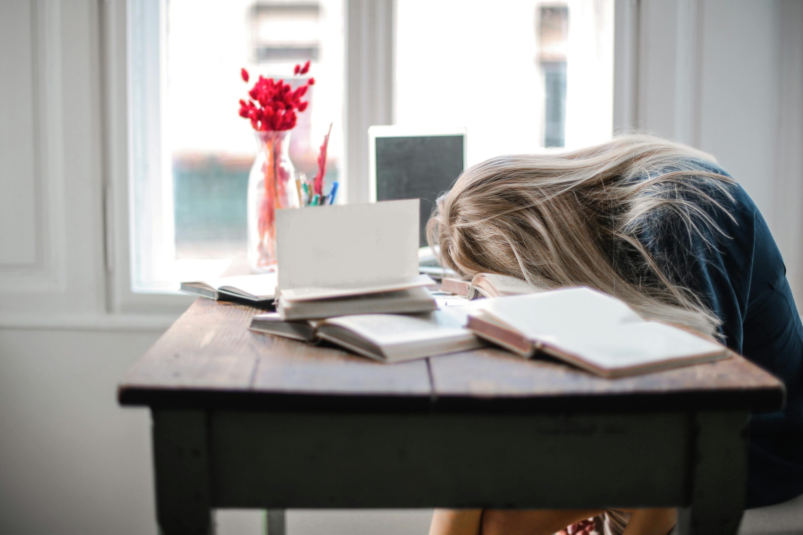 Person with long blonde hair rests head on desk with books flowers and laptop